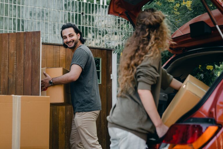 Cheerful modern male and female in comfortable casual clothes unpacking car trunk in green garden of new house while moving in together